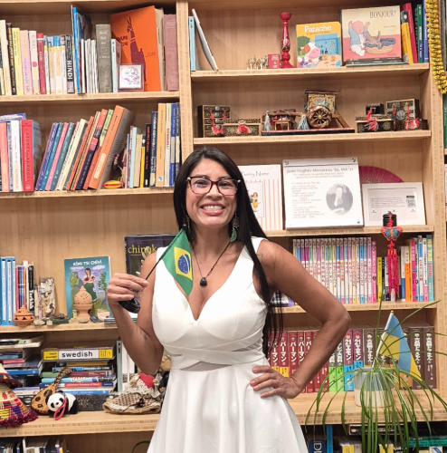 A woman in a white dress in front of a bookshelf holding a Brazilian flag. flag. 