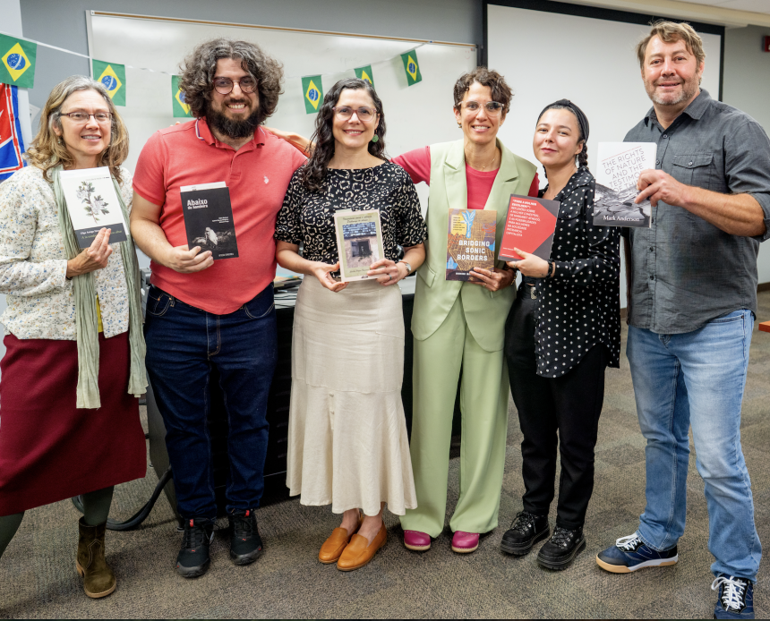 Six people stand in front of a wall decorated with Brazilian flags, each holding a book.