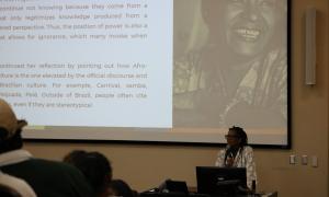 Djamila Ribeiro holds a microphone. Behind her there is an image of Lelia Gonzalez